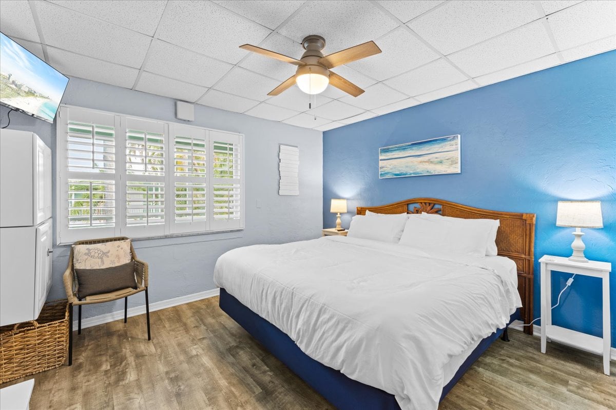 Spacious bedroom with a king bed, smart TV, ceiling fan, and built-in storage — a relaxing retreat with bright windows and beachy artwork.