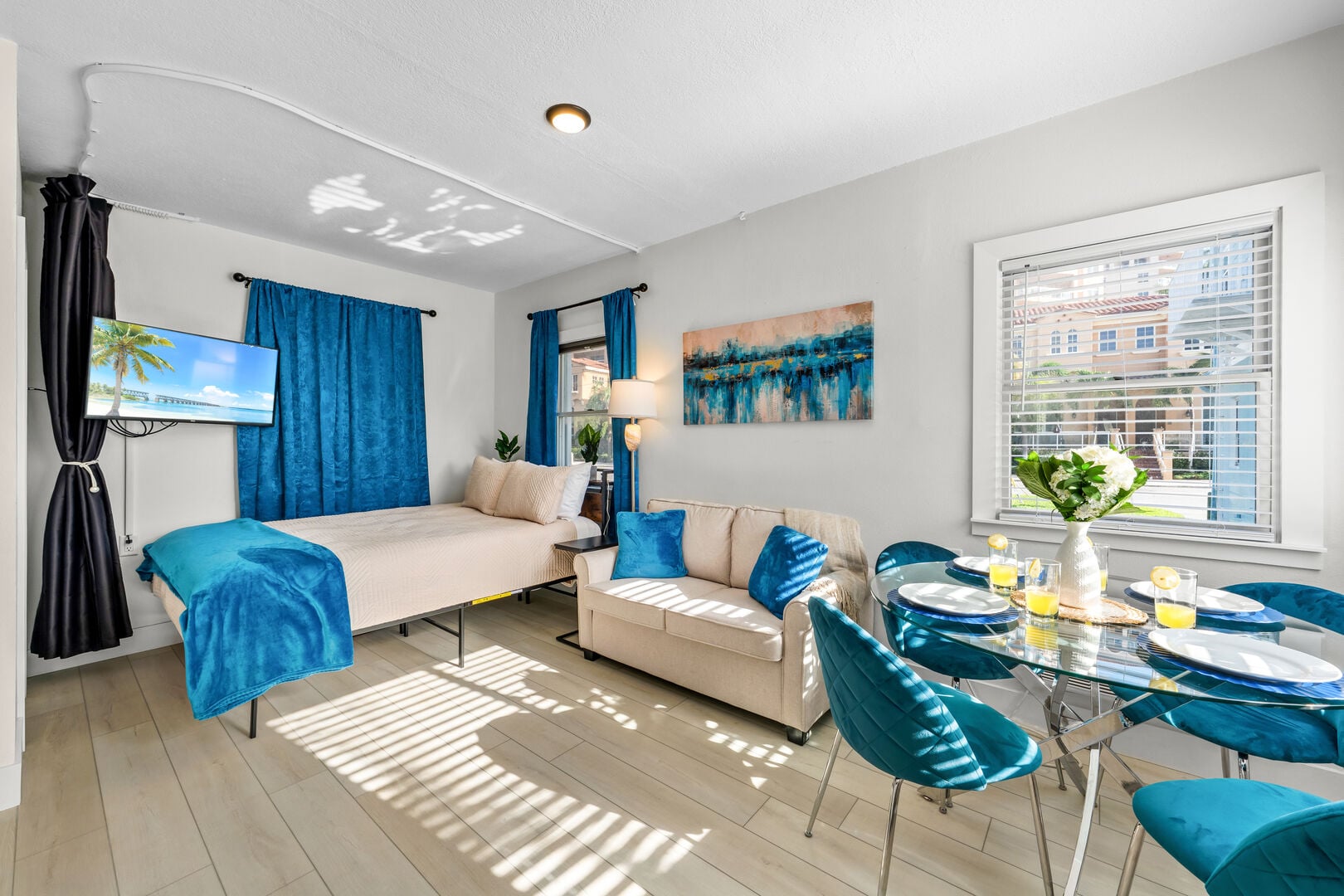 The full open-plan interior of Coral Resort E2: coastal-blue decor, wall-mounted smart TV, a cushioned daybed sleeping area (with a privacy curtain), plush loveseat and a glass dining table for four — all filled with Florida sunshine.